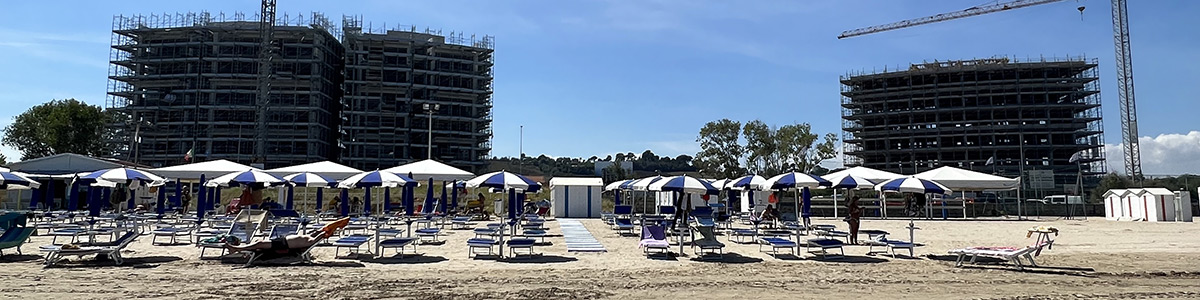 Foto scattata dalla battigia sulla spiaggia antistante. In primo piano si vedono gli ombrelloni e subito dietro i due enormi edifici in costruzione