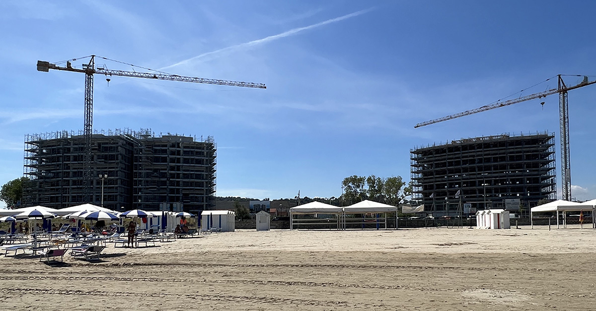 Foto scattata dalla battigia sulla spiaggia antistante. In primo piano si vedono gli ombrelloni e subito dietro i due enormi edifici in costruzione