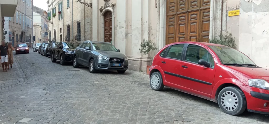 7 auto parcheggiate lungo via San Martino e un'auto che passa. A lato due pedoni si stringono verso il muro per farla passare