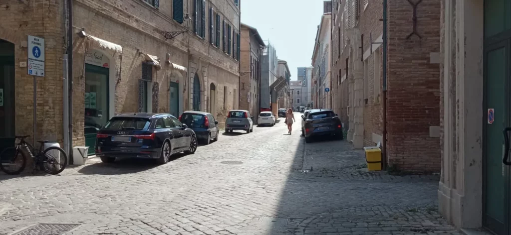 8 auto parcheggiate lungo via Fratelli Bandiera. Un. pedone cammina al centro della strada perché i lati sono occupati