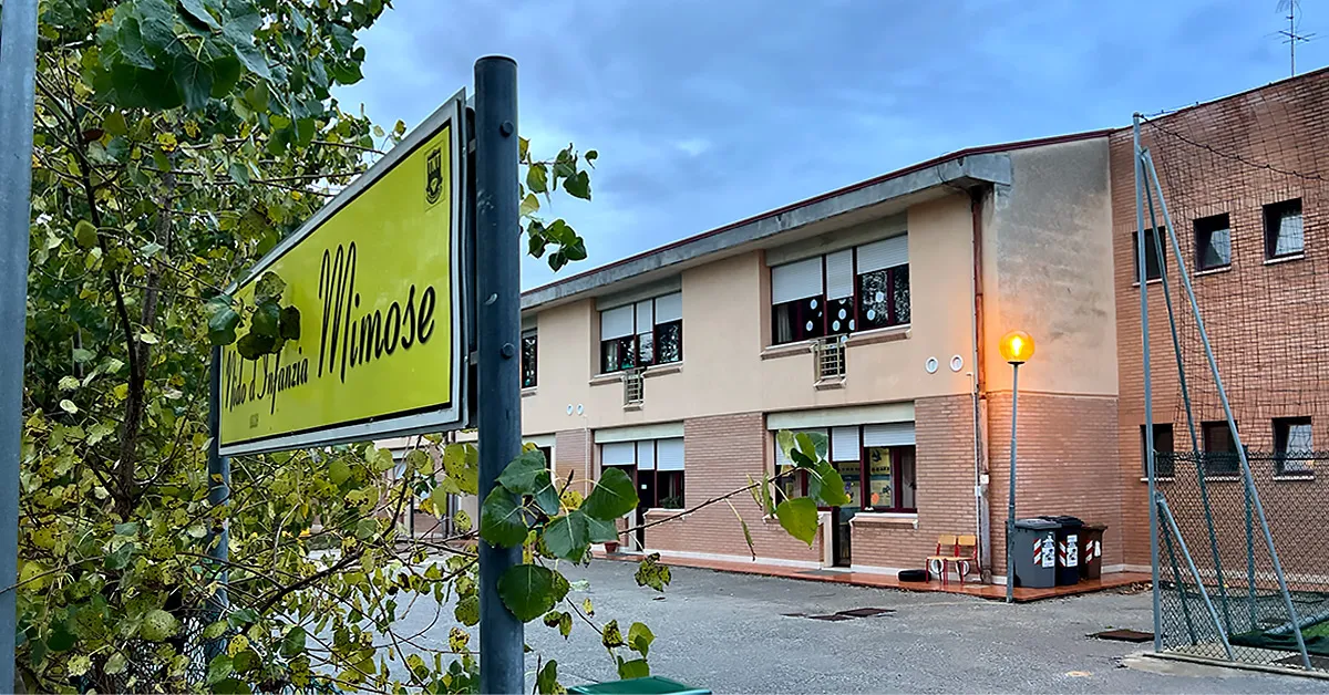 L'edificio dell'ex-asilo Mimose fotografato dall'esterno del recinto. In primo piano sulla sinistra si legge il cartello giallo "Nido d'Infanzia Mimose". L'edificio ha un piano terra e un primo piano ed è a ridosso della scuola Rodari. È in buone condizioni, costruito con mattoncini rossi, ma è dismesso.