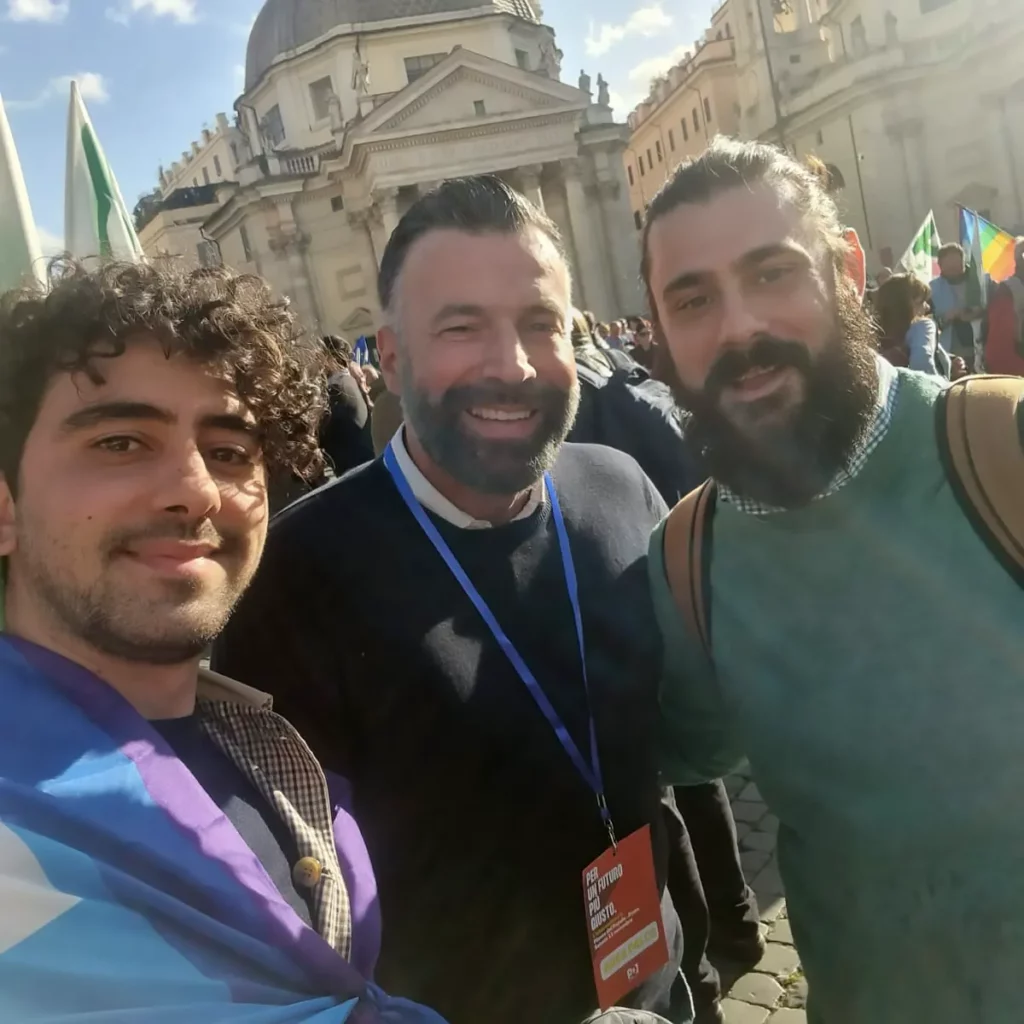 Selfie scattato da Giovanni, che sulle spalle ha la bandiera della pace. Zan è al centro, barba corta curata, sorridente e con maglione nero